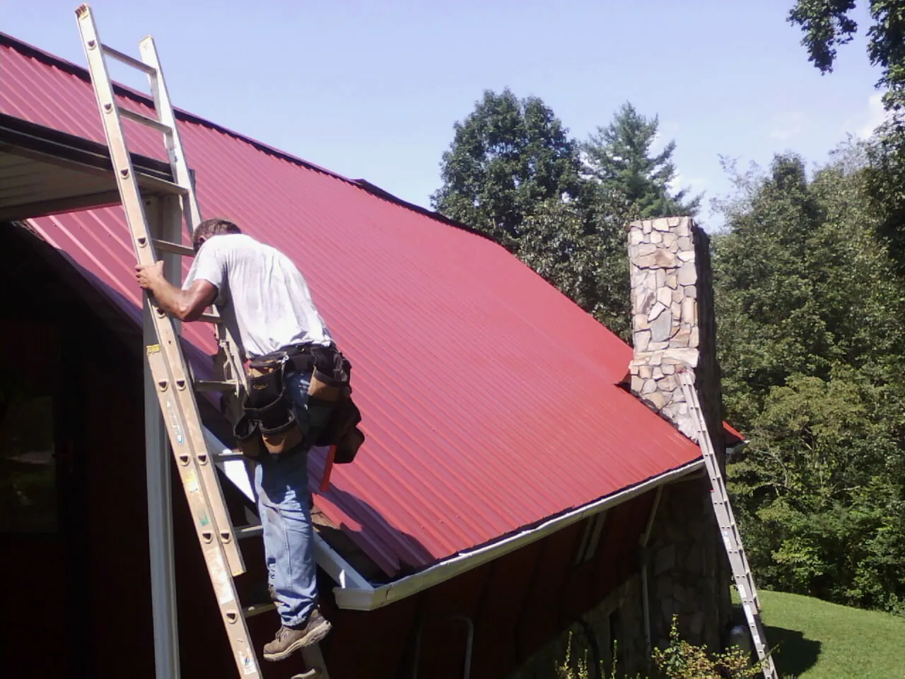 Roofer on a ladder beside a red metal roof and stone chimney — Soffit & Fascia Repair by Roofing Brothers in Montana Mines